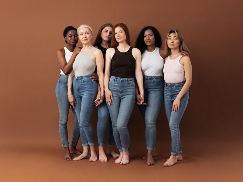 Sechs Frauen unterschiedlichen Alters und Ethnie posieren in Jeans und Top vor einer braunen Fotoleinwand.