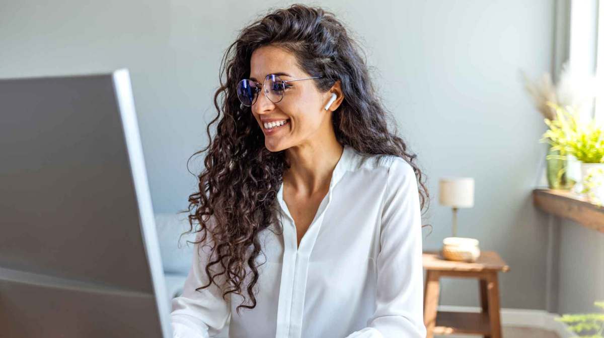 Junge Frau mit dunklen Locken sitzt am Schreibtisch mit Kopfhörern und spricht in einem Online-Meeting mit jemandem.