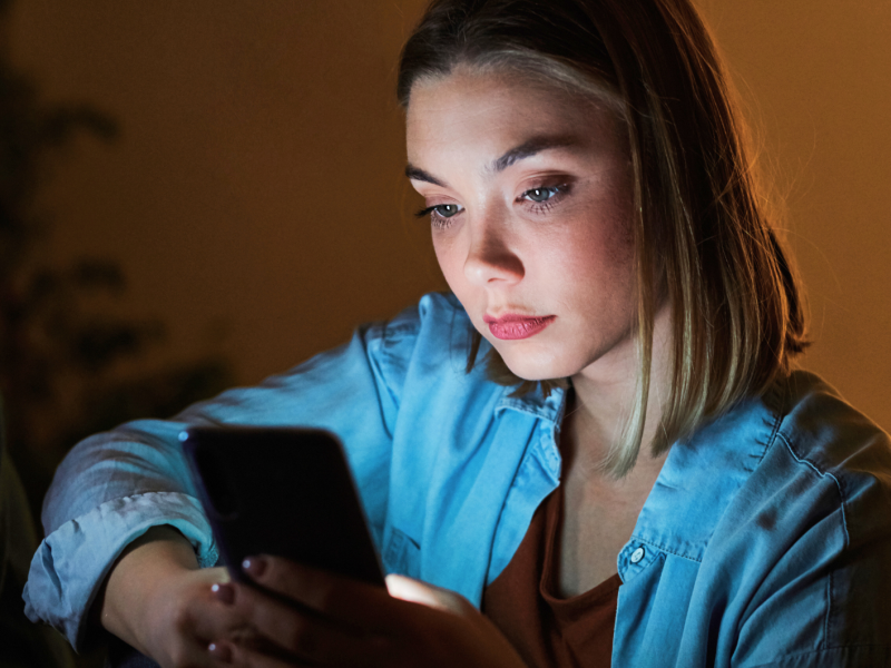 Eine junge Frau mit ernstem Gesichtsausdruck liest auf ihrem Handy, umgeben von gedämpftem, orangefarbenem Licht.