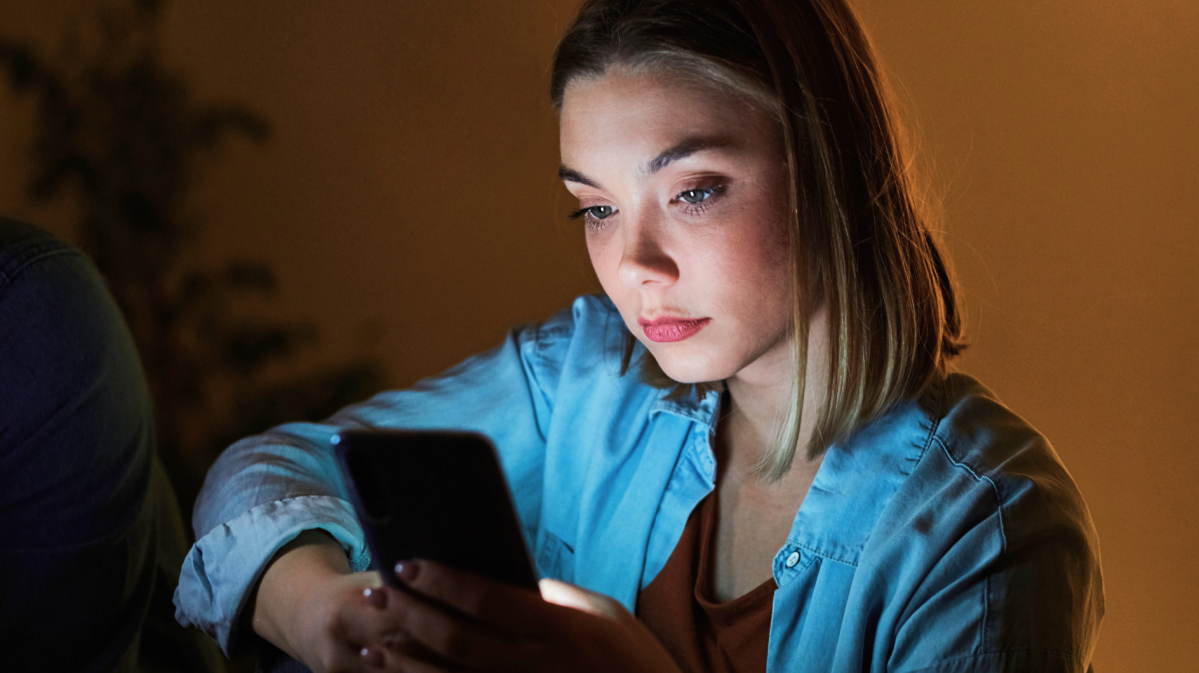 Eine junge Frau mit ernstem Gesichtsausdruck liest auf ihrem Handy, umgeben von gedämpftem, orangefarbenem Licht.