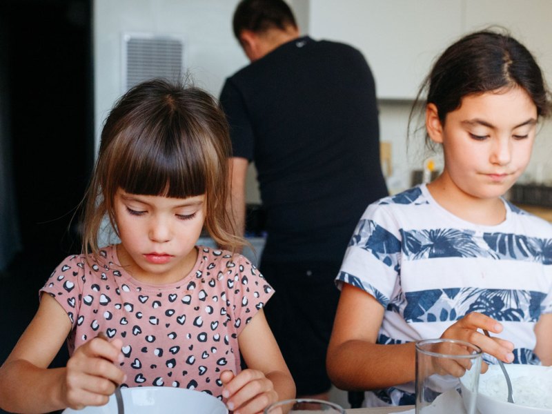 Schwestern sitzen am Tisch und sind beide auf ihre Schüssel mit Essen konzentriert. Im Hintergrund sieht man den Vater kochen.