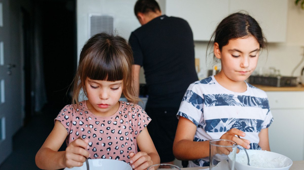 Schwestern sitzen am Tisch und sind beide auf ihre Schüssel mit Essen konzentriert. Im Hintergrund sieht man den Vater kochen.