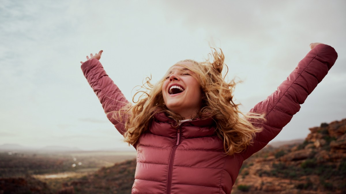 Frau mit rosa WInterjacke, die die Arme in die Luft schwingt und lacht, während sie draußen auf einem Berg steht