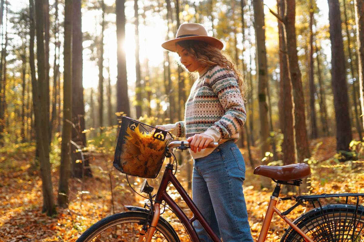 Lächelnde junge Frau in einem Hut und einem stilvollen Pullover und einem Fahrrad, genießt das Herbstwetter im Wald, unter den gelben Blättern.