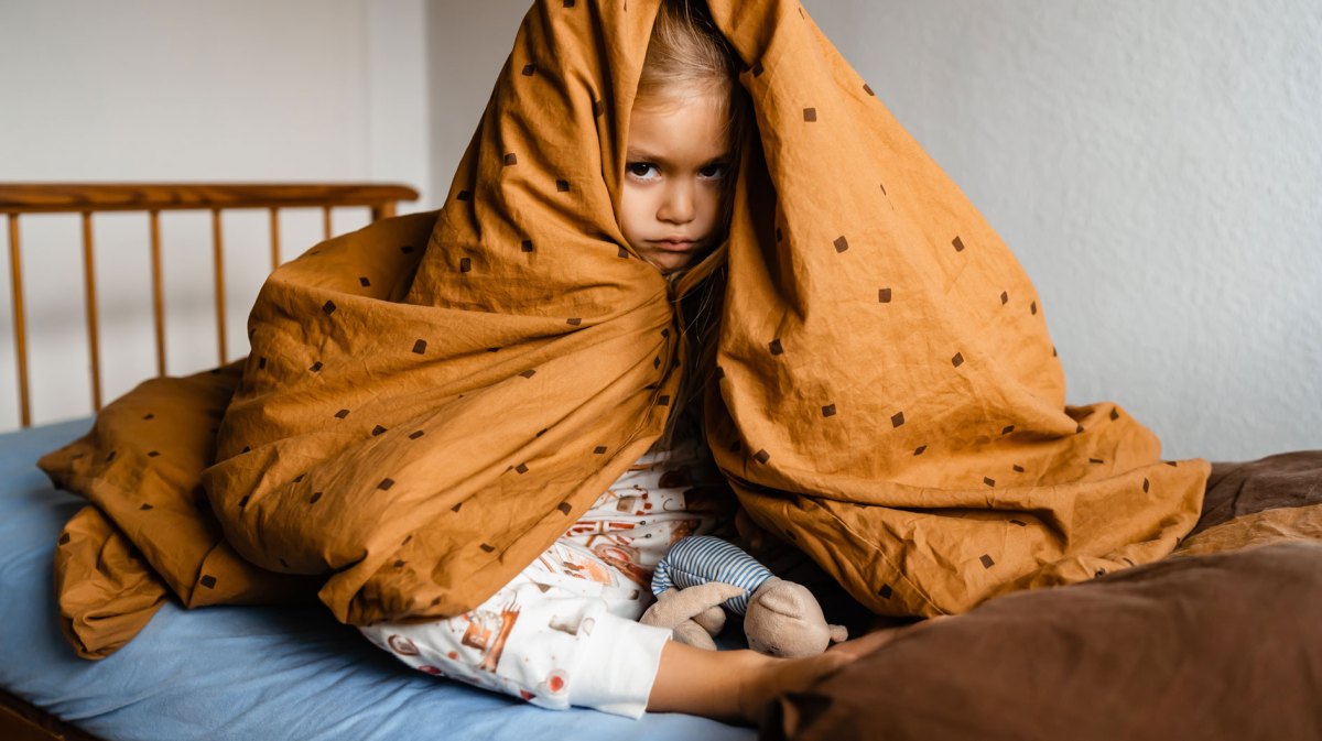 Kind, ca 5 Jahre, sitzt versteckt unter seiner Decke auf dem Bett und schaut verärgert in die Kamera.