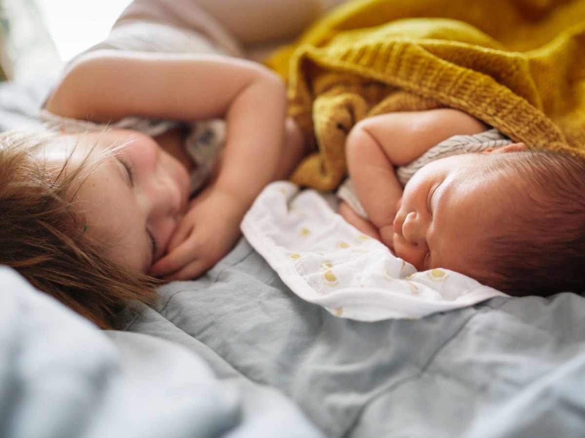 Ältere Schwester im Kleinkindalter liegt auf dem Bett neben ihrem neugeborenen Geschwisterchen.