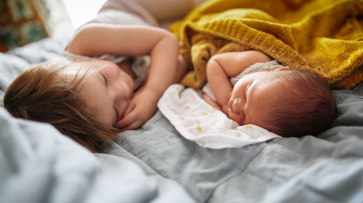 Ältere Schwester im Kleinkindalter liegt auf dem Bett neben ihrem neugeborenen Geschwisterchen.