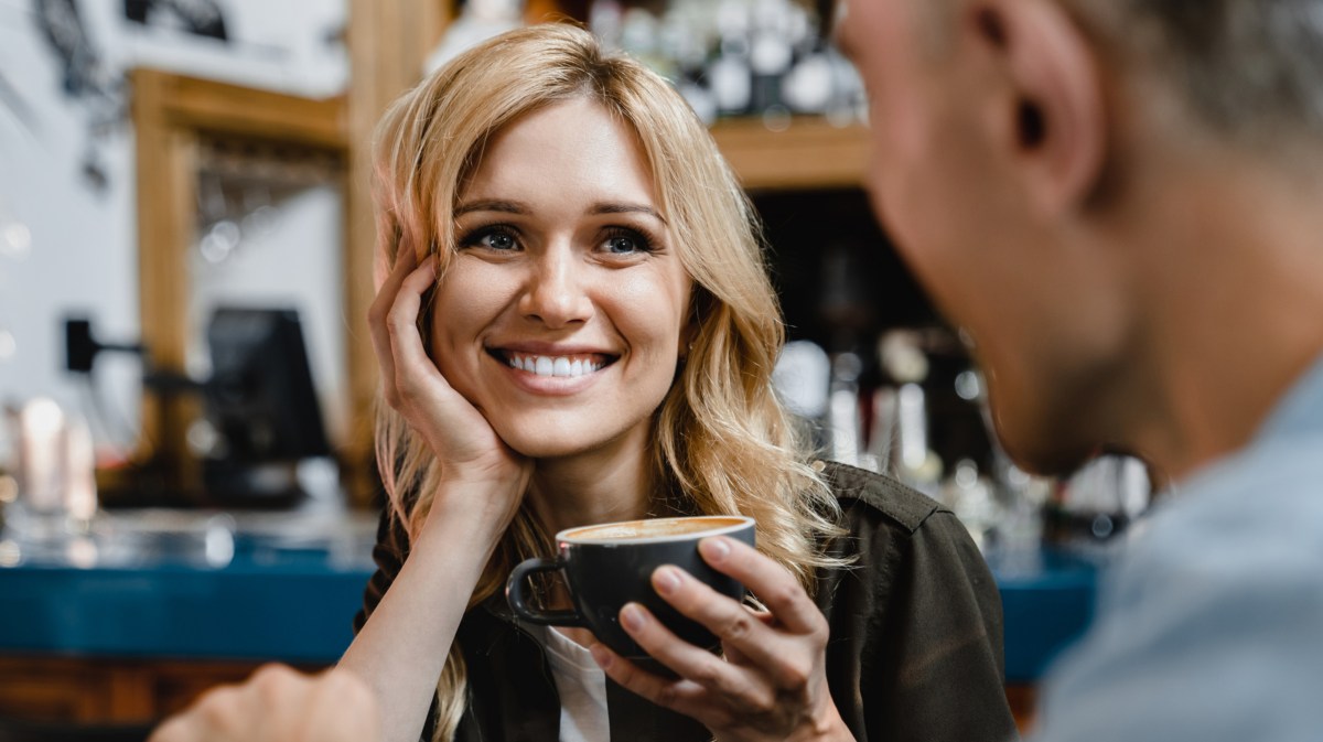 Blonde Frau hält eine Kaffeetasse in der Hand und sieht ihr Gegenüber verliebt an.