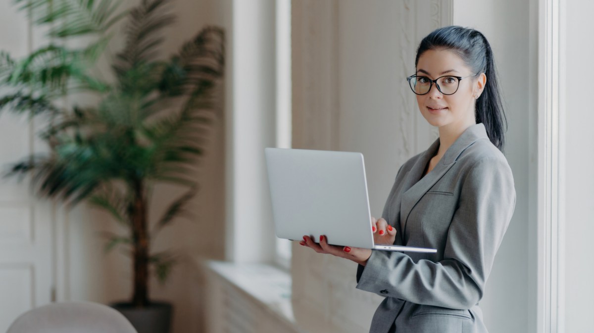 Frau mit Laptop steht auf der Arbeit an einer Wand gelehnt.