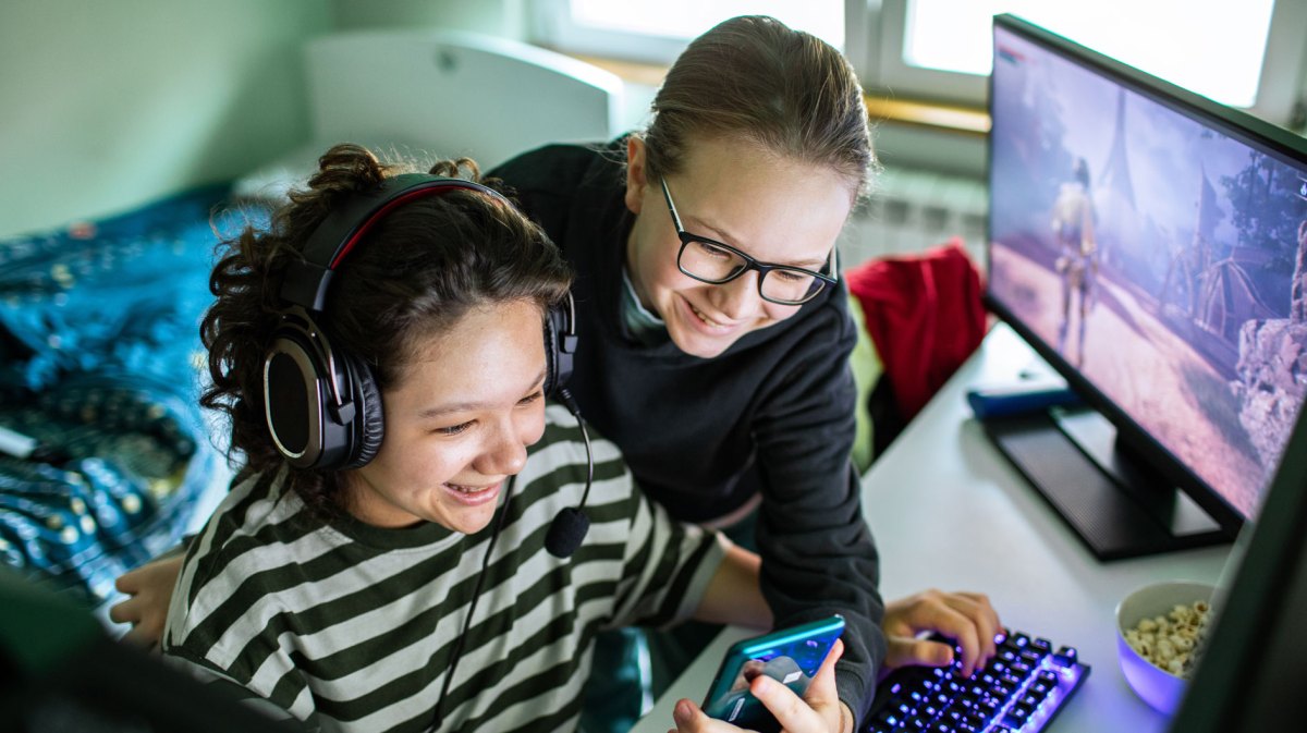 Zwei Schwestern im Kinderzimmer, eine sitzt mit Kopfhörern vorm PC und zockt, die andere zeigt ihr gerade etwas auf ihrem Handy.