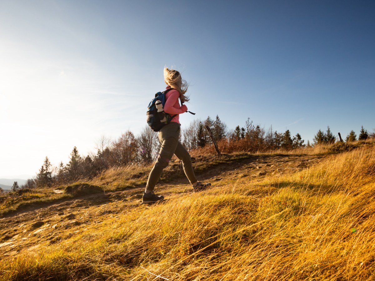 Frau auf einem Berg mit Wiese, die mit Rucksack und Winterjacke hochgeht