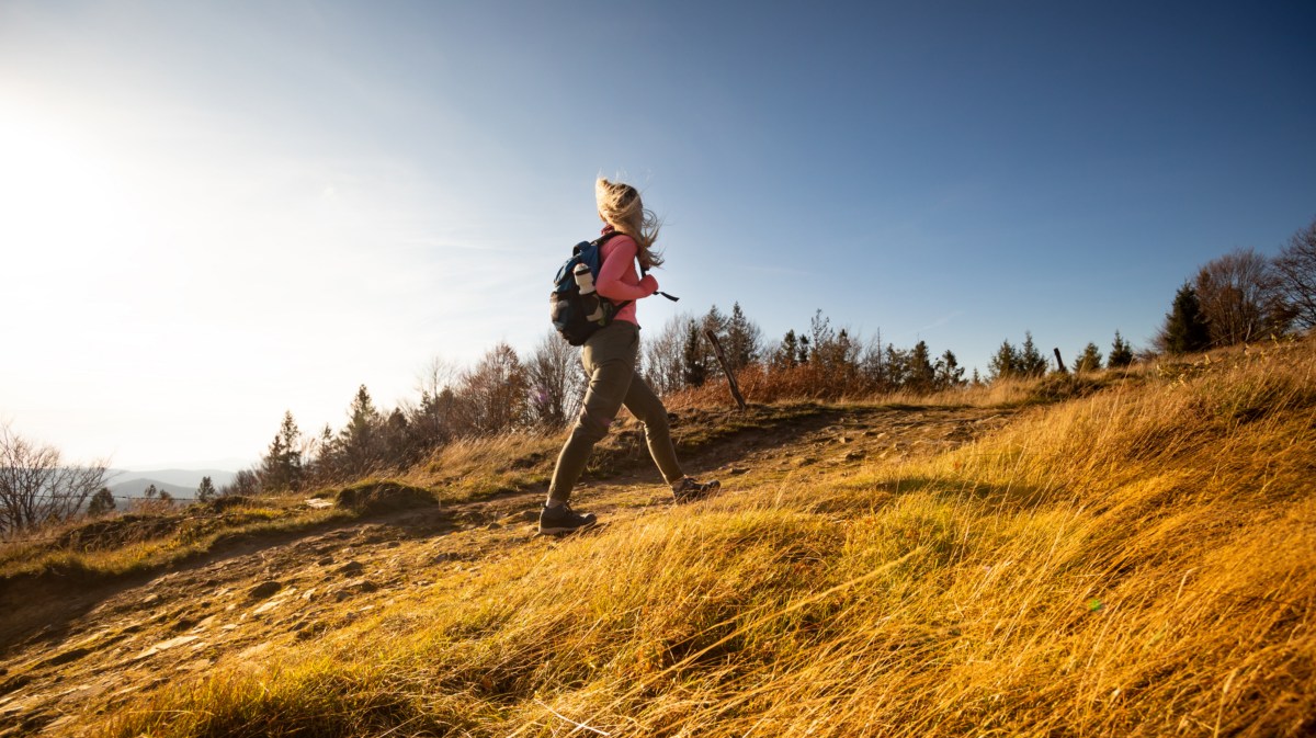 Frau auf einem Berg mit Wiese, die mit Rucksack und Winterjacke hochgeht