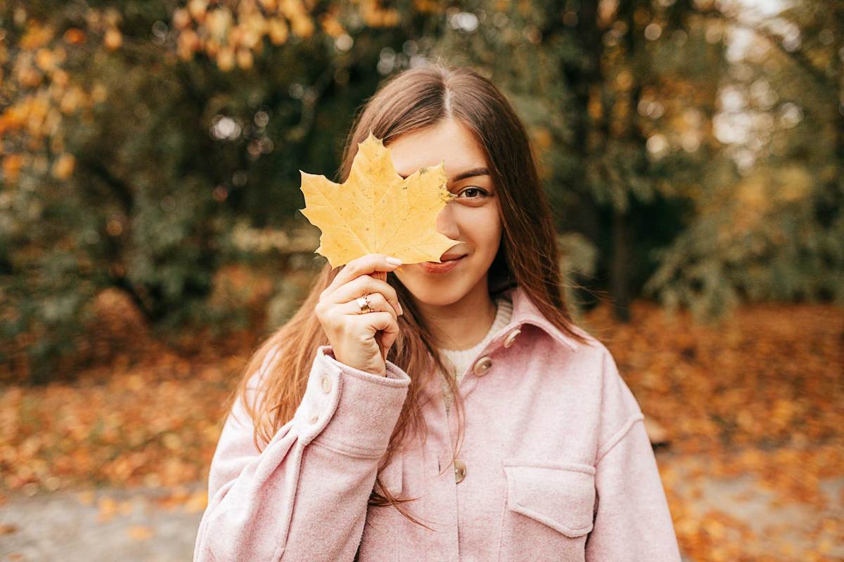 Positive braunhaarige Frau mit langen glatten Haaren in gemütlicher Herbstkleidung hat Spaß vor der Kamera, posiert mit Blatt auf ihrem Gesicht im Herbstpark. Altweibersommer.