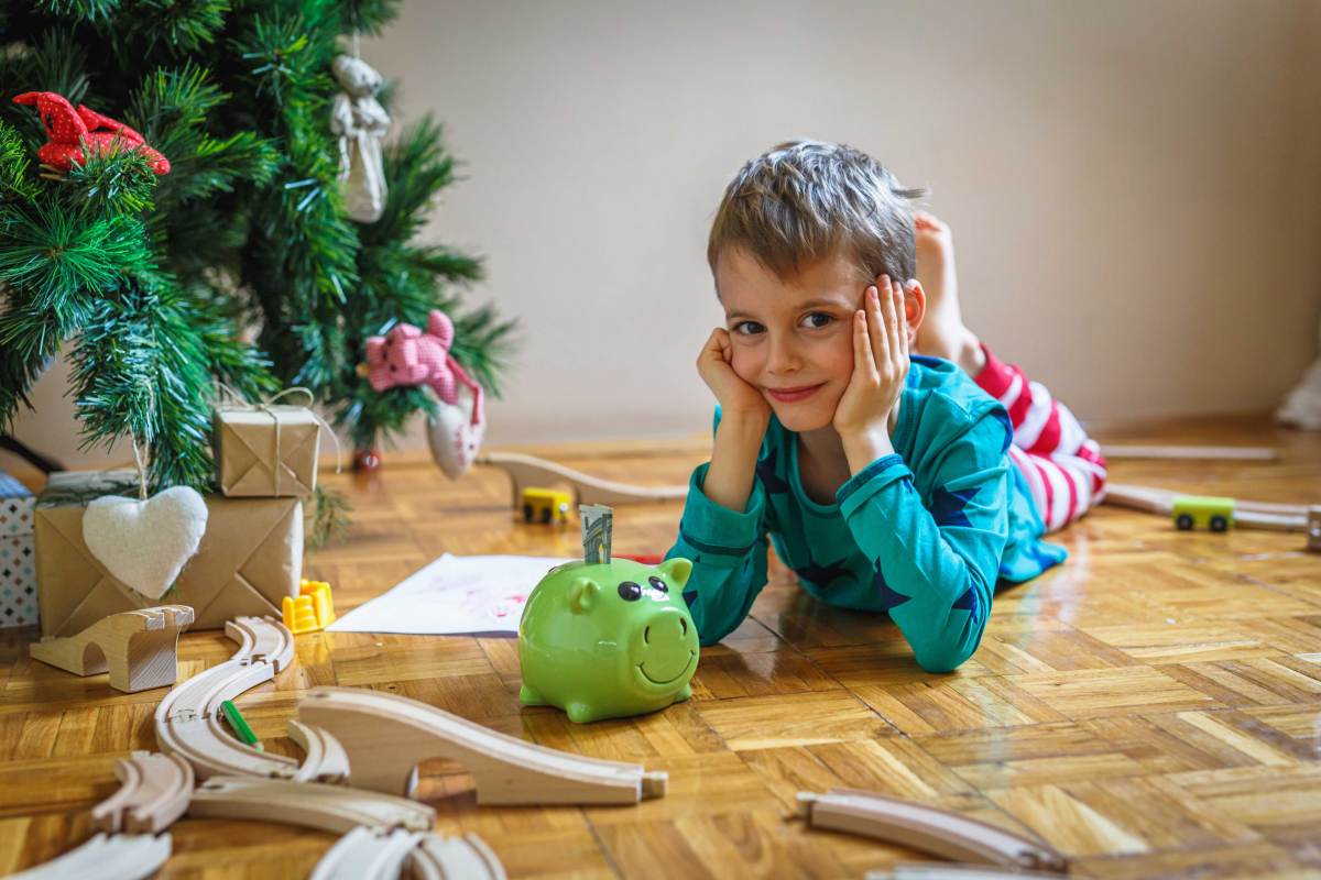 Kleiner Junge liegt neben dem Weihnachtsbaum, mit einem Sparschwein vor sich, in dem ein Geldschein steckt.