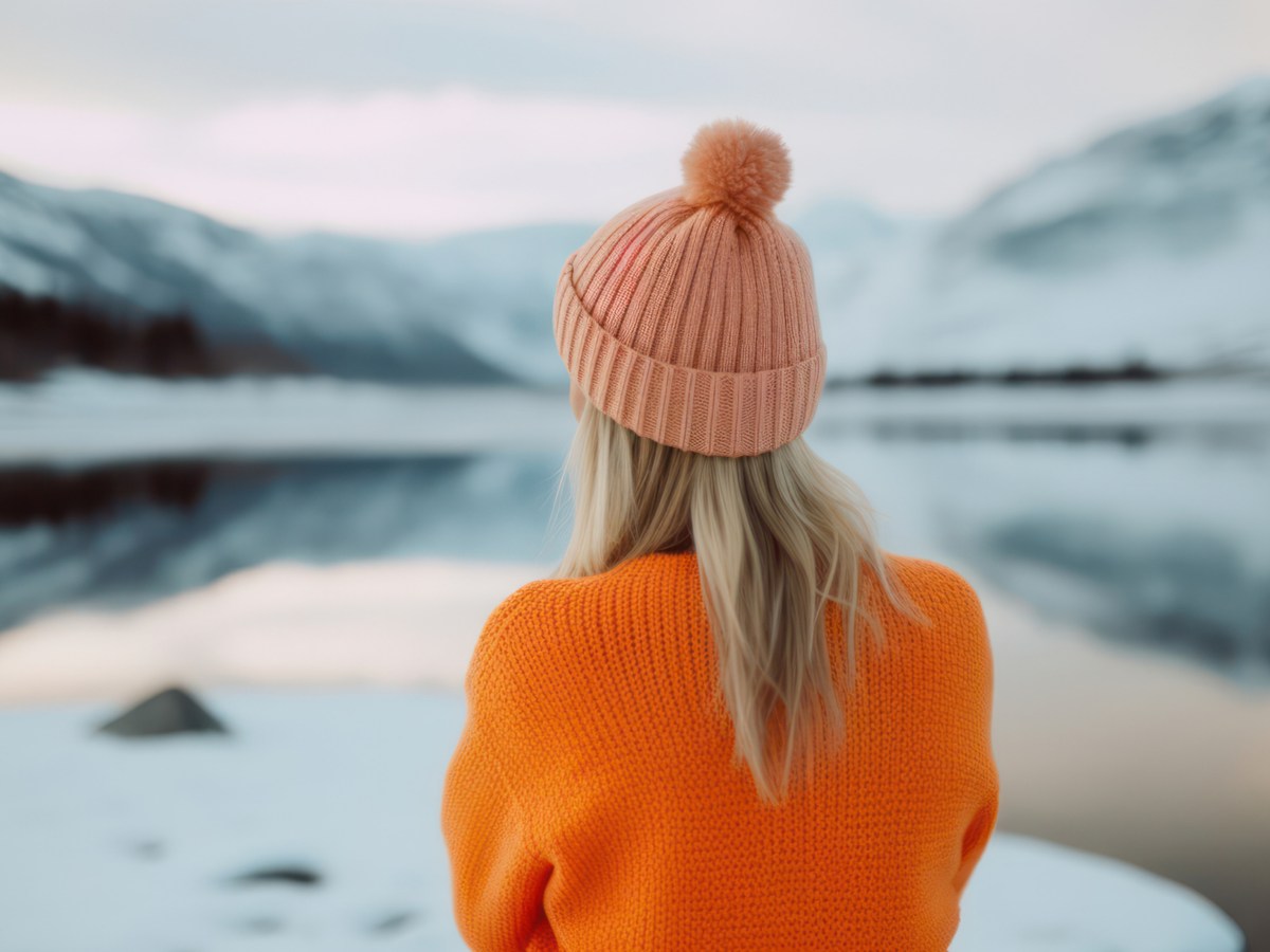 Blonde Frau mit Mütze auf dem Kopf, die vor einem See und einer Schneelandschaft steht