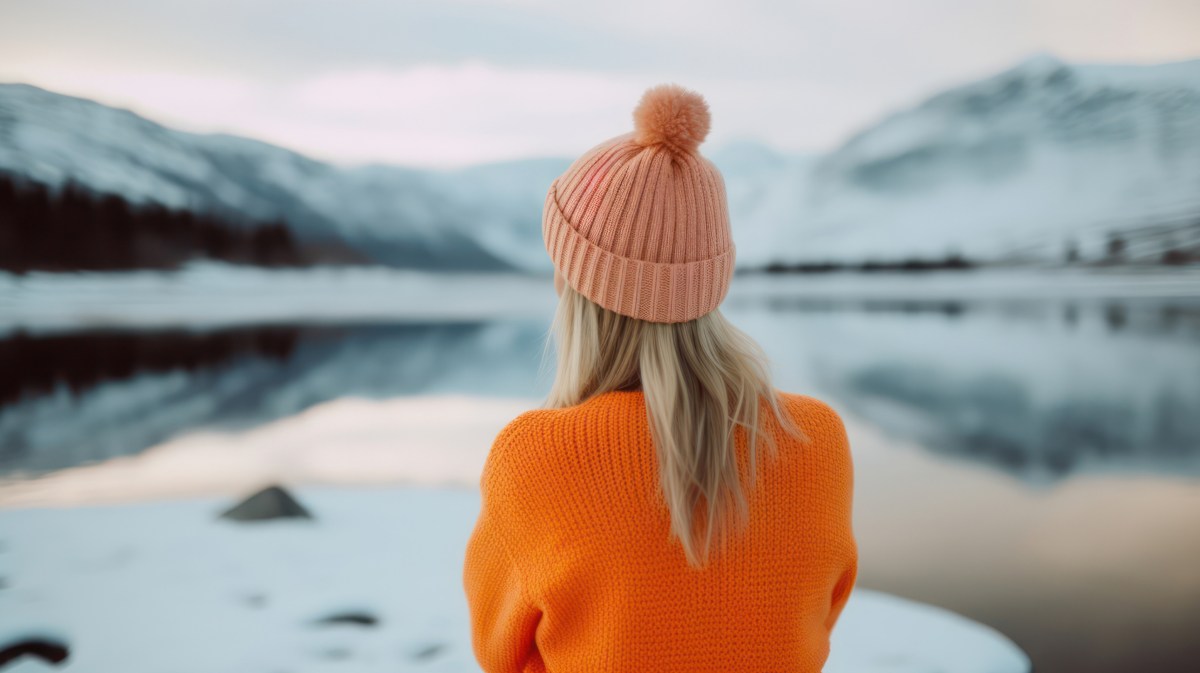 Blonde Frau mit Mütze auf dem Kopf, die vor einem See und einer Schneelandschaft steht