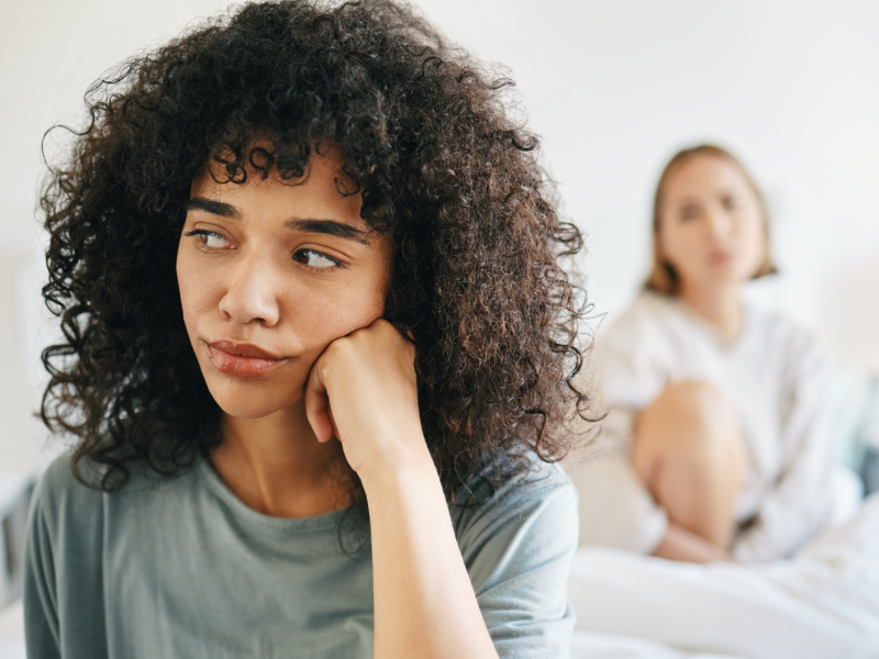 Frau, die nachdenklich zur Seite schaut, während im Hintergrund eine andere Frau auf sie blickt und auf einem Bett sitzt.