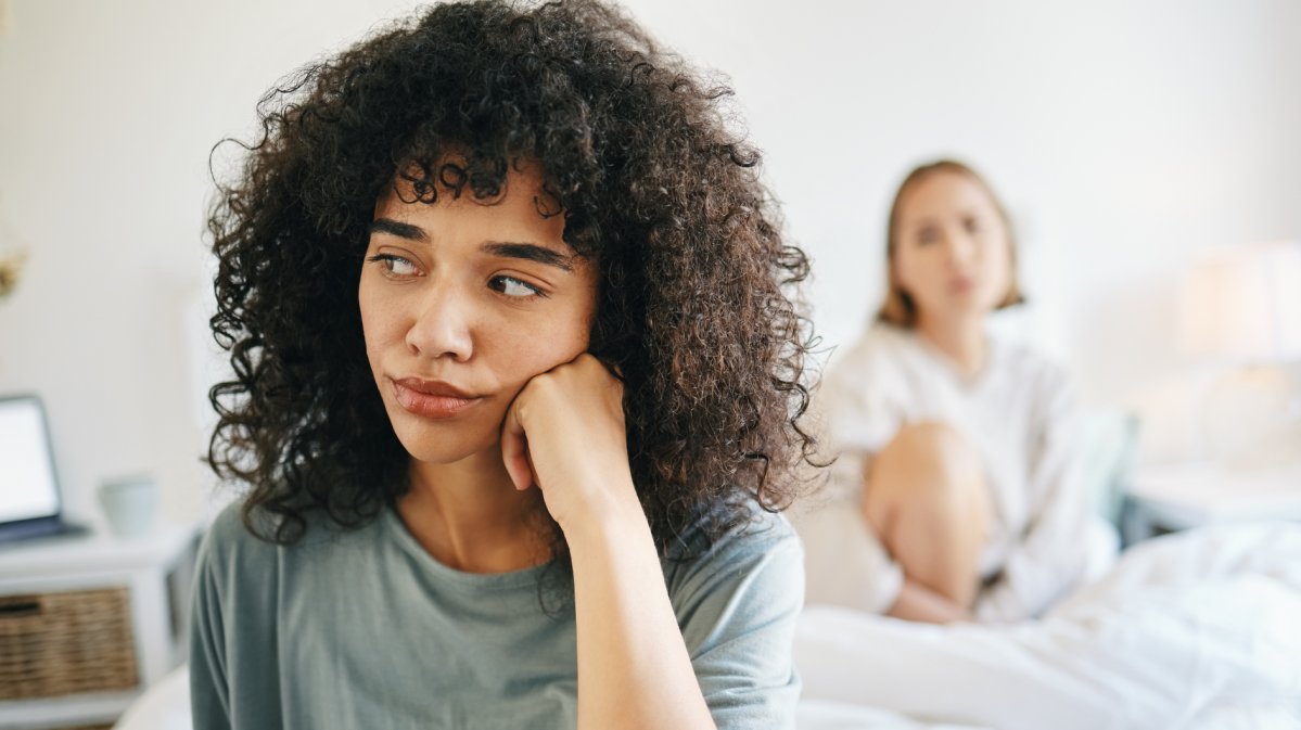 Frau, die nachdenklich zur Seite schaut, während im Hintergrund eine andere Frau auf sie blickt und auf einem Bett sitzt.