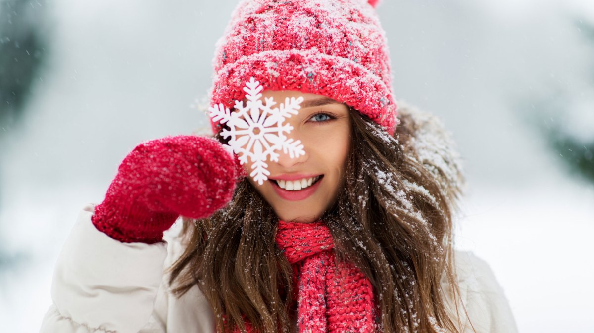 Frau in Winterbekleidung im Schnee hält sich Schneeflocke vor das Gesicht und lächelt in die Kamera