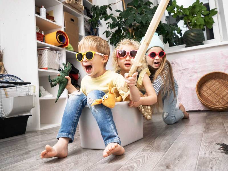 Drei Kinder spielen drinnen. Zwei sitzen mit Sonnenbrillen und Spielzeug in den Händen in einer Plastikbox, die vom dritten Kind durch den Raum geschoben wird.