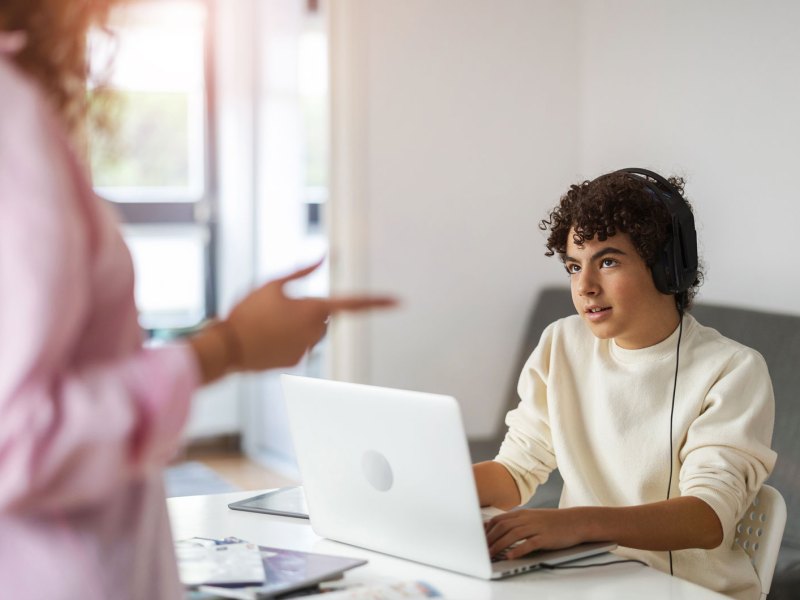 Teenager-Sohn sitzt am Laptop und spielt, seine Mutter ist nicht glücklich darüber.