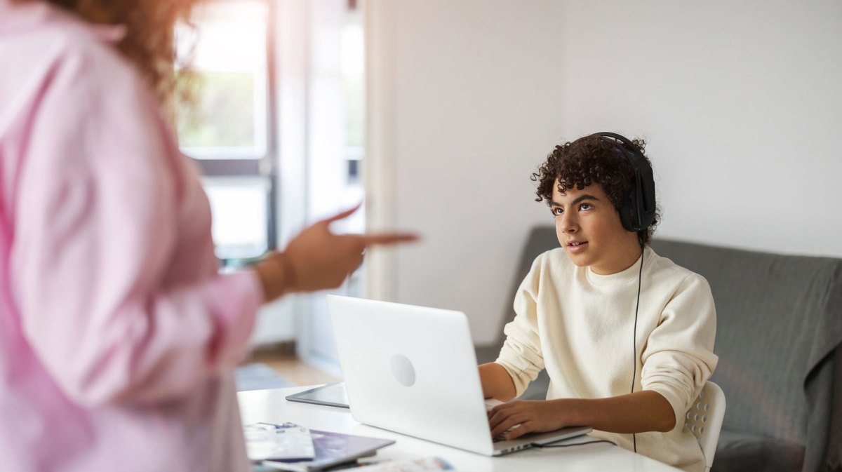 Teenager-Sohn sitzt am Laptop und spielt, seine Mutter ist nicht glücklich darüber.