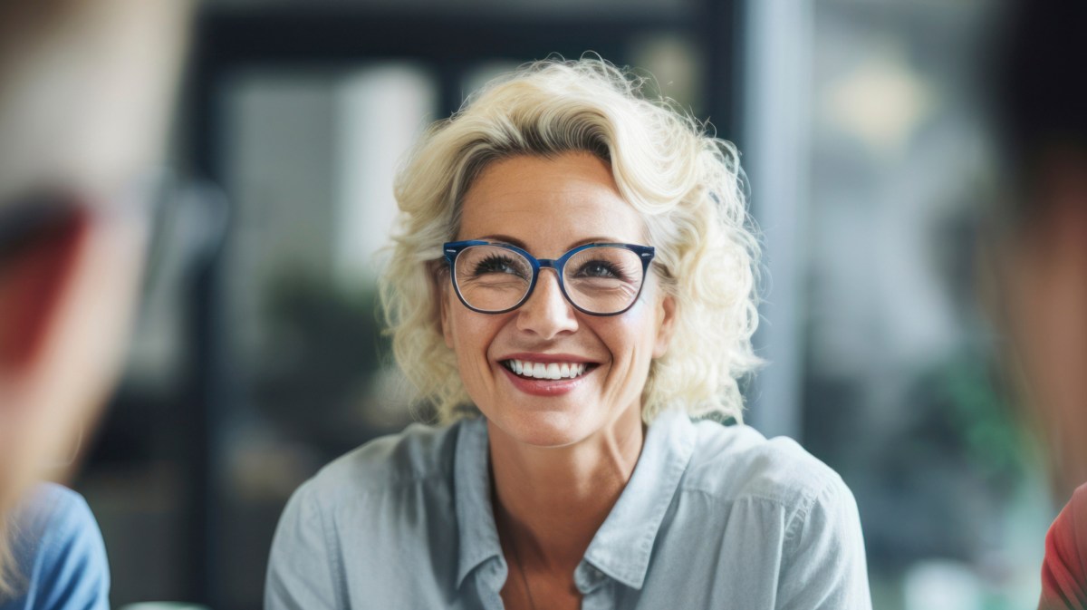 Frau mittleren Alters mit Brille und blonden kurzen Locken lächelt auf der Arbeit.