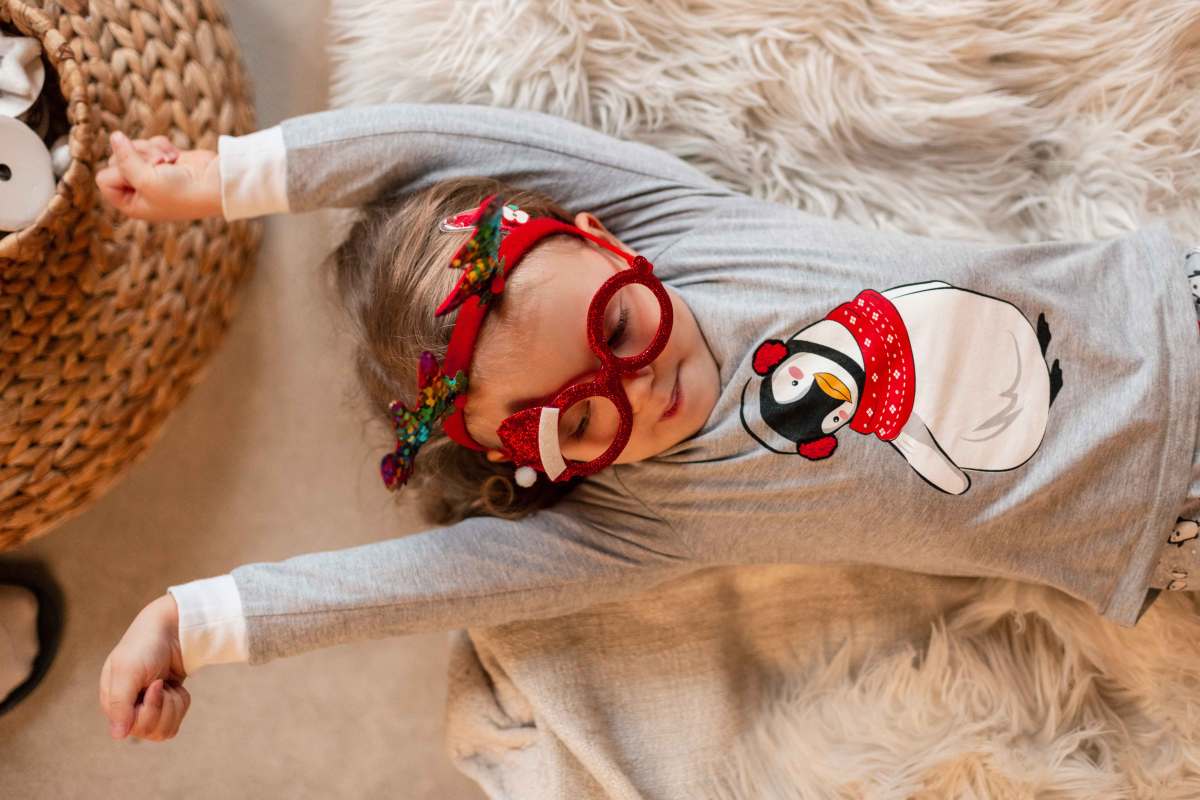 Kleines Mädchen Liegt in Weihnachtspulli und mit Partybrille auf einem Teppich und streckt sich vor Müdigkeit.