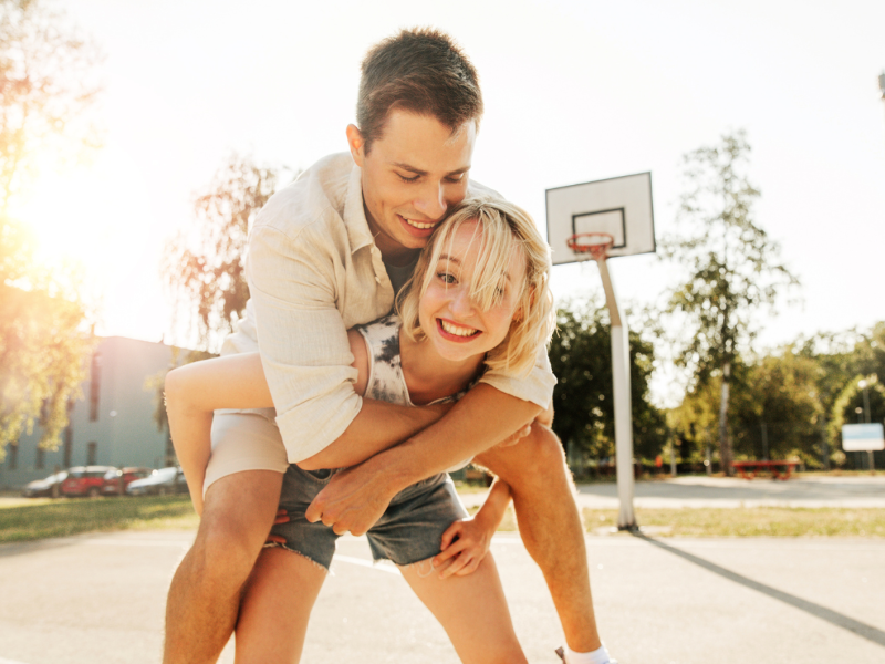 Frau und Mann auf einem Basketballplatz und die Frau den Mann Huckepack nimmt.