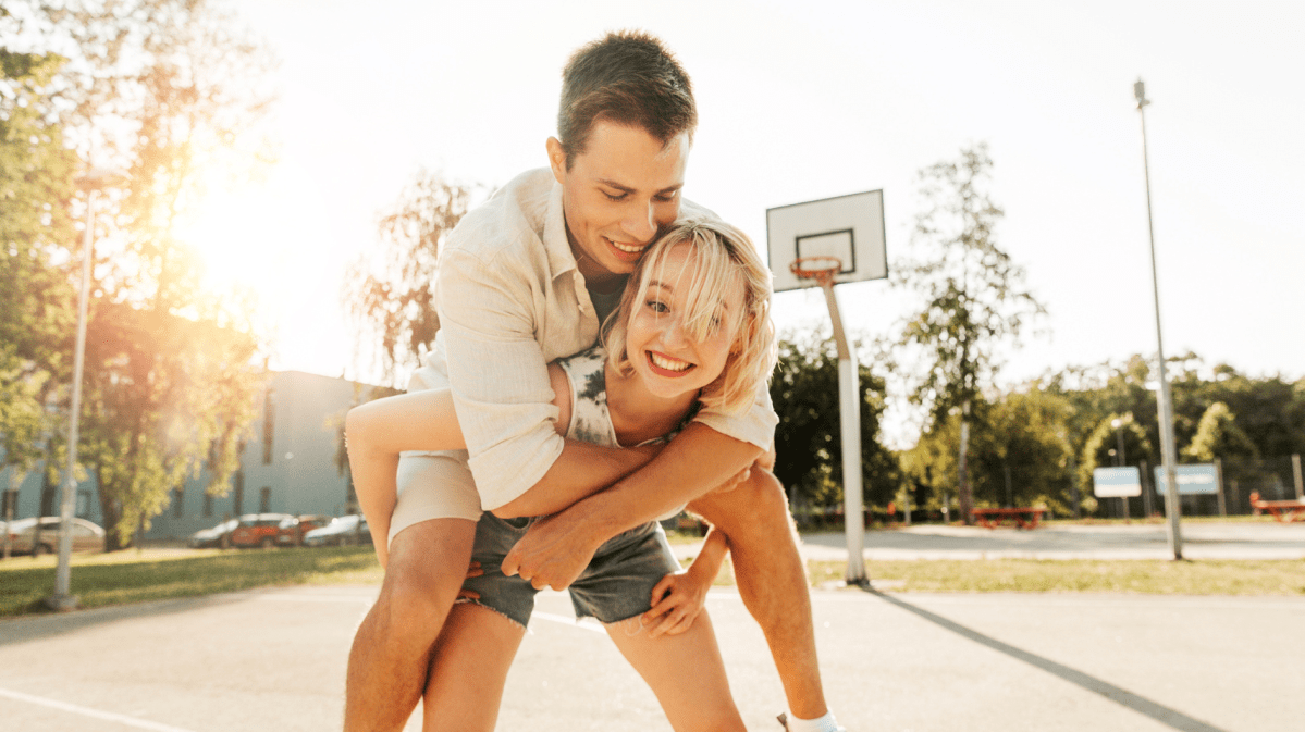 Frau und Mann auf einem Basketballplatz und die Frau den Mann Huckepack nimmt.