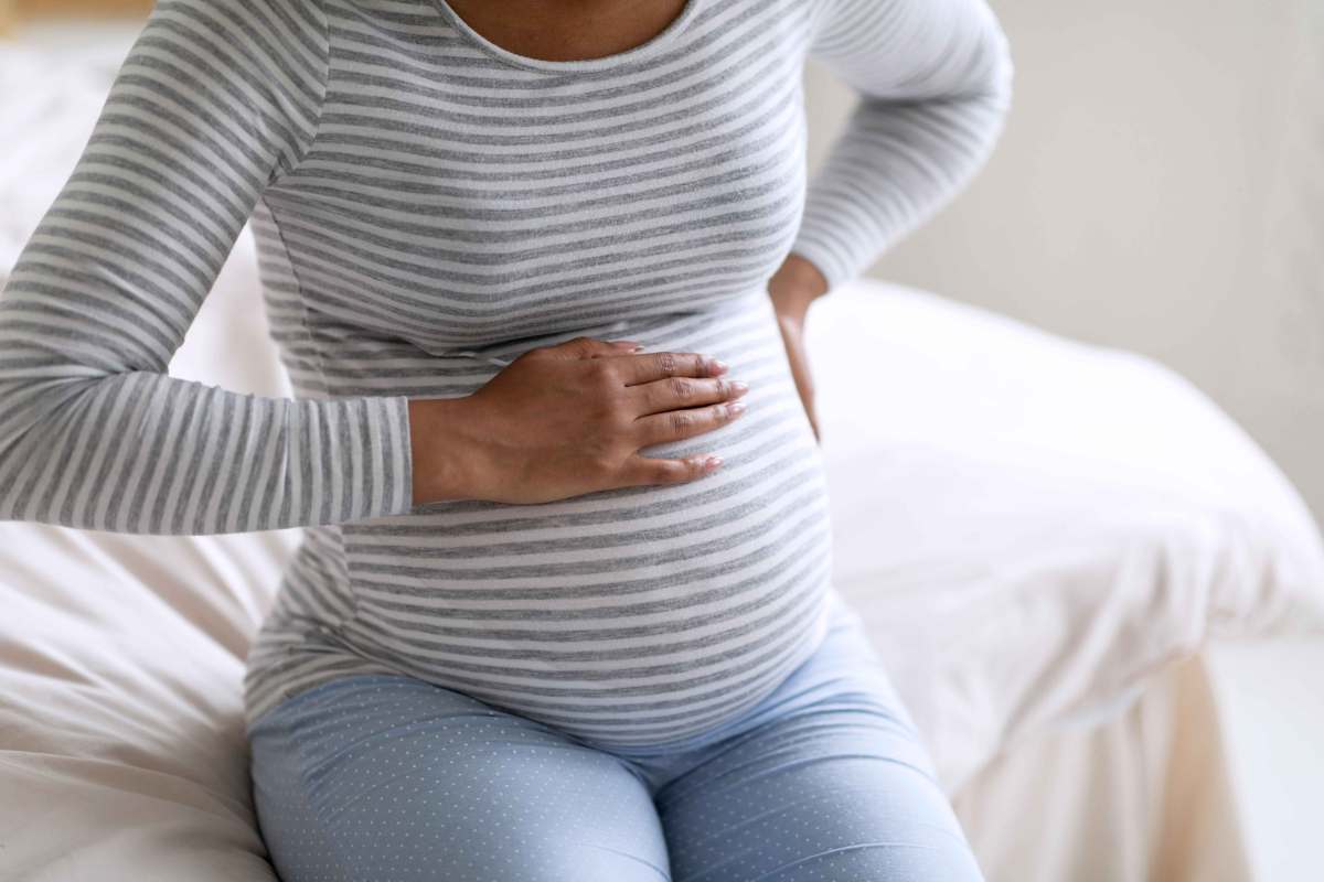 Schwangere Frau sitzt auf dem Bett und hält sich sowohl den Bauch als auch den Rücken.