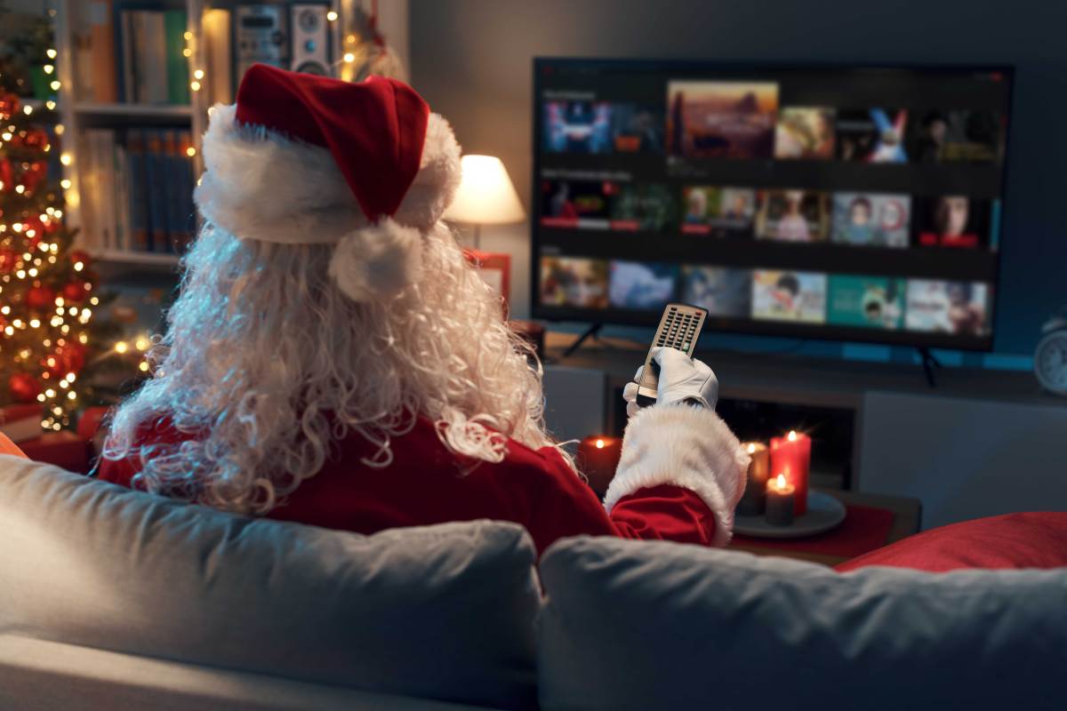 Weihnachtsmann sitzt mit dem Rücken zur Kamera auf der Couch vor einem Fernseher und überlegt, was er schauen kann. Neben ihm steht ein Weihnachtsbaum.