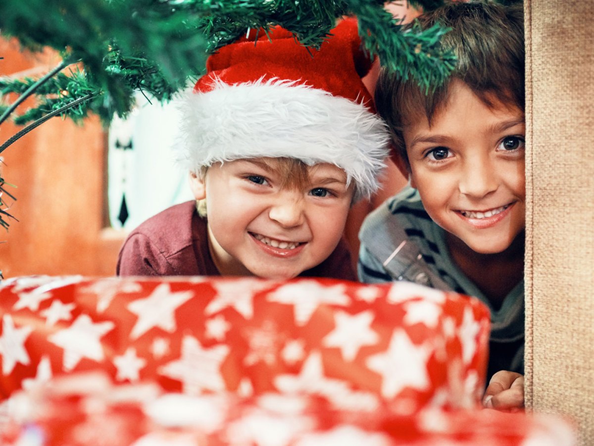 Zwei Jungs schauen neugierig unter dem Tannenbaum hindurch zu Geschenken.