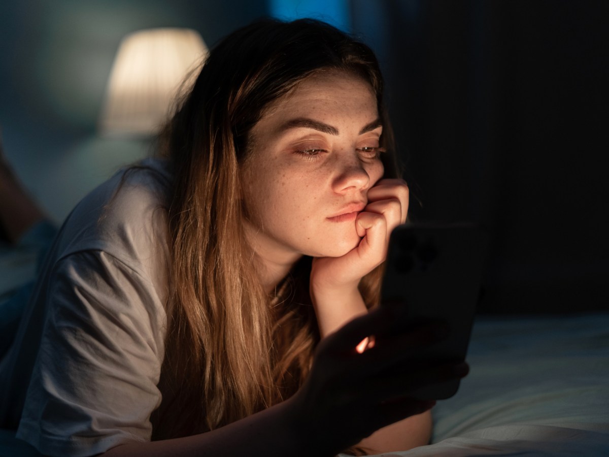 Eine Frau liegt im Bett und schaut gelangweilt auf ihr Handy.