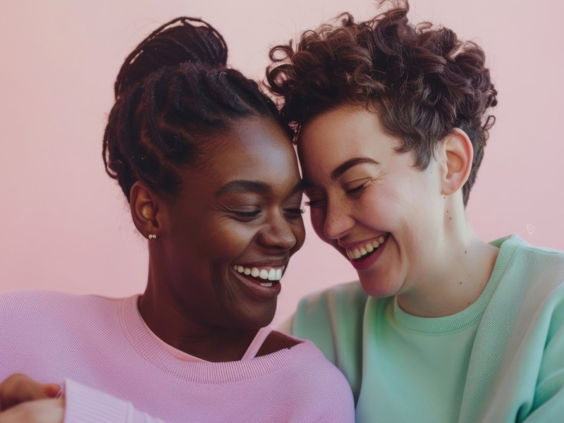 Zwei Frauen vor einer rosa Wand, die ihre Köpfe aneinander lehnen und lachen.