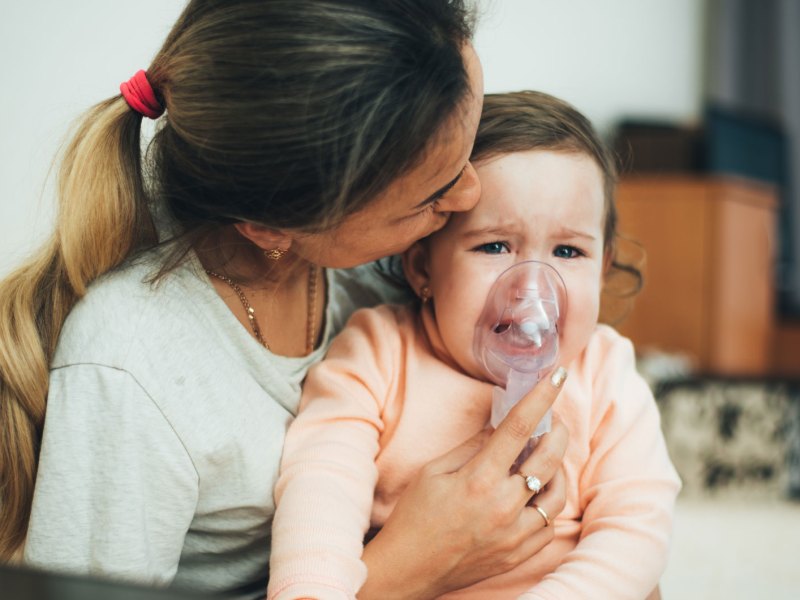 Mutter hält ihrer Tochter eine Atemmaske eines Inhaliergeräts vor Nase und Mund.