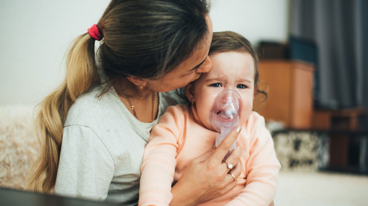 Mutter hält ihrer Tochter eine Atemmaske eines Inhaliergeräts vor Nase und Mund.