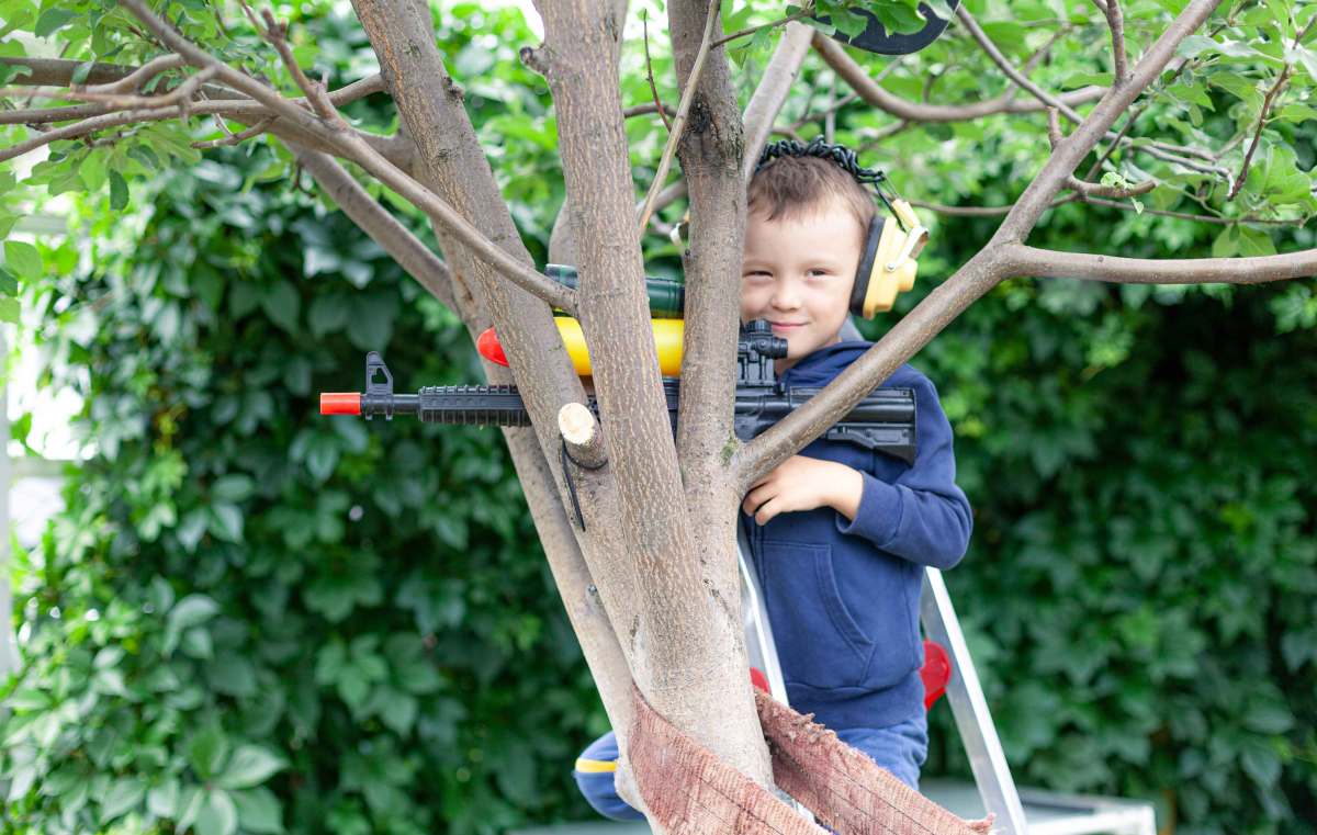 Kleiner Junge sitzt mit Ohrenschützern und einem Spielzeuggewehr in einem Baum und schaut verschmitzt in die Kamera.