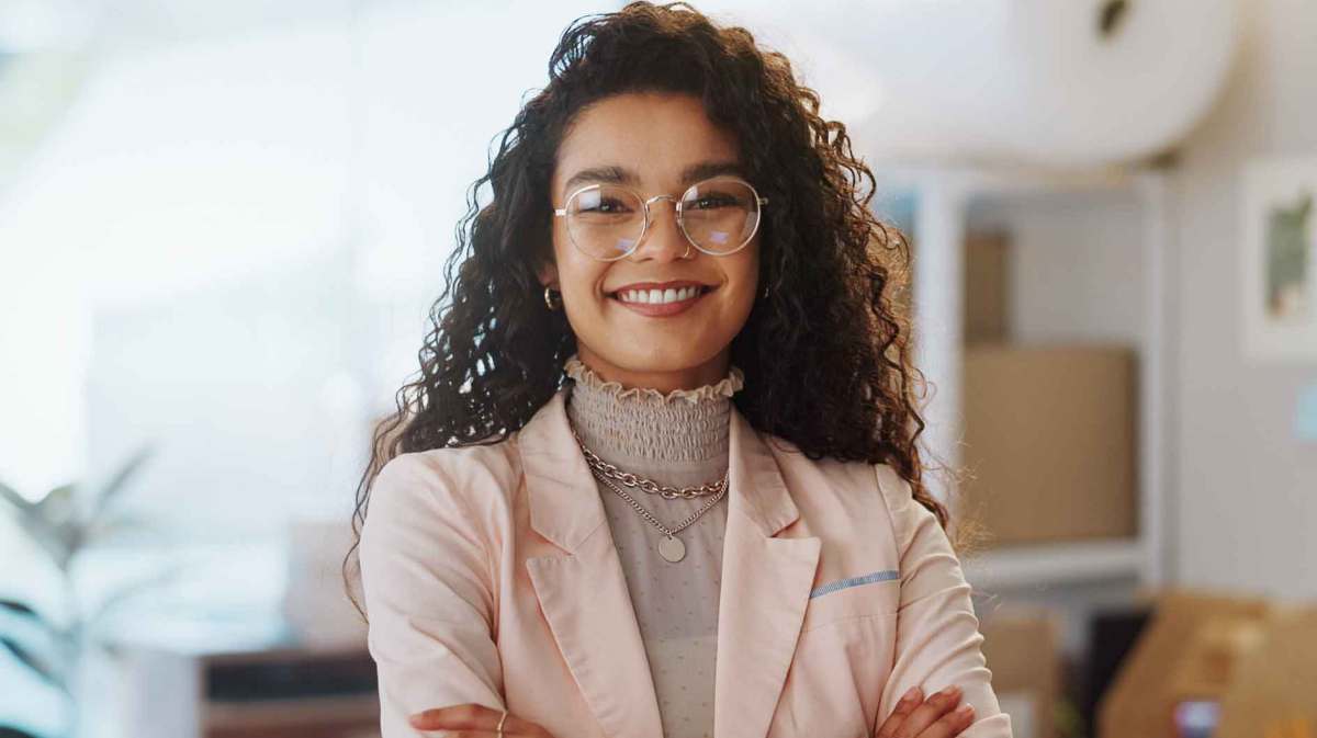 Junge Frau mit Locken und Brille lächelt selbstbewusst in die Kamera. Sie trägt ein Business-Outfit und steht auf der Arbeit.