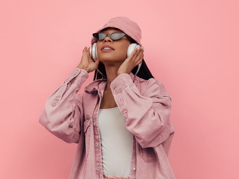 Frau vor rosa Wand mit Kopfhörern. Hut und Sonnenbrille auf dem Kopf.