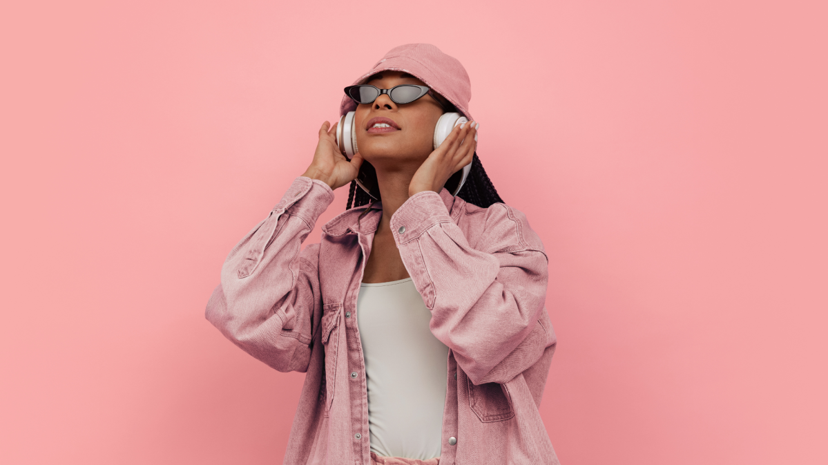 Frau vor rosa Wand mit Kopfhörern. Hut und Sonnenbrille auf dem Kopf.