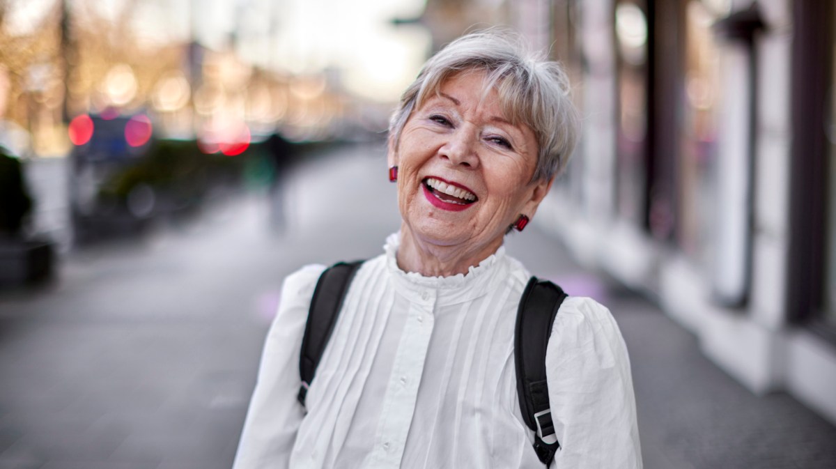 Modern gekleidete, ältere Frau mit einer weißen Bluse, roten Ohrringen und weißen Haaren lacht in die Kamera.