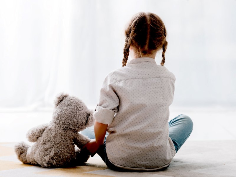 Mädchen, ca. 8 Jahre, sitzt mit dem Rücke zur Kamera und hält einen Teddybären an der Pfote fest.