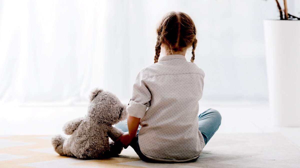 Mädchen, ca. 8 Jahre, sitzt mit dem Rücke zur Kamera und hält einen Teddybären an der Pfote fest.