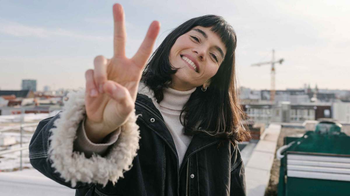 Junge Frau mit braunen Haaren zeigt das Peace Zeichen.