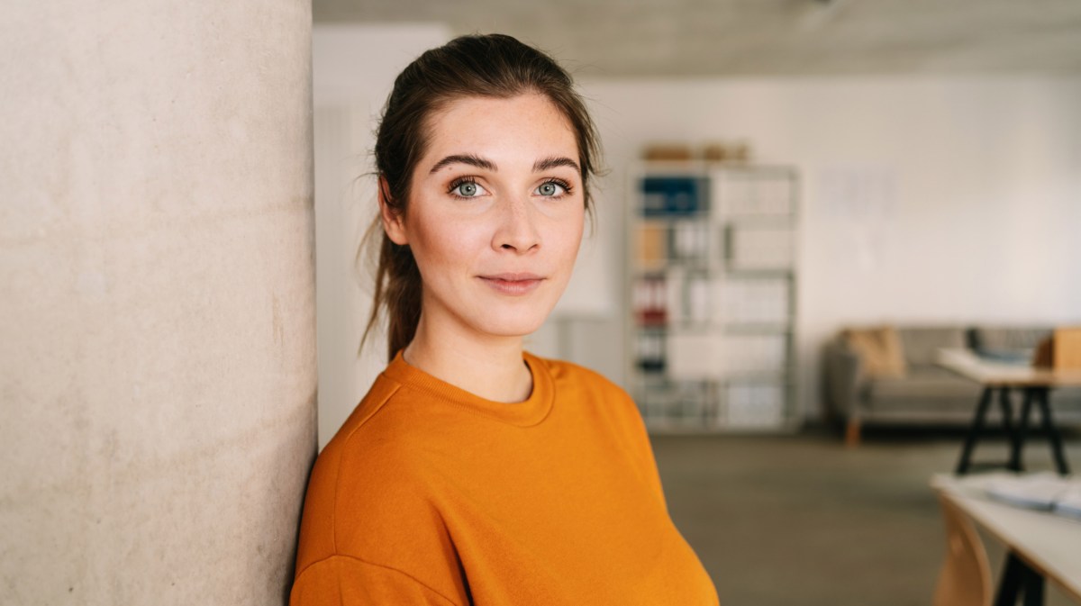 Junge Frau in orangefarbenen Pullover schaut direkt in Kamera.