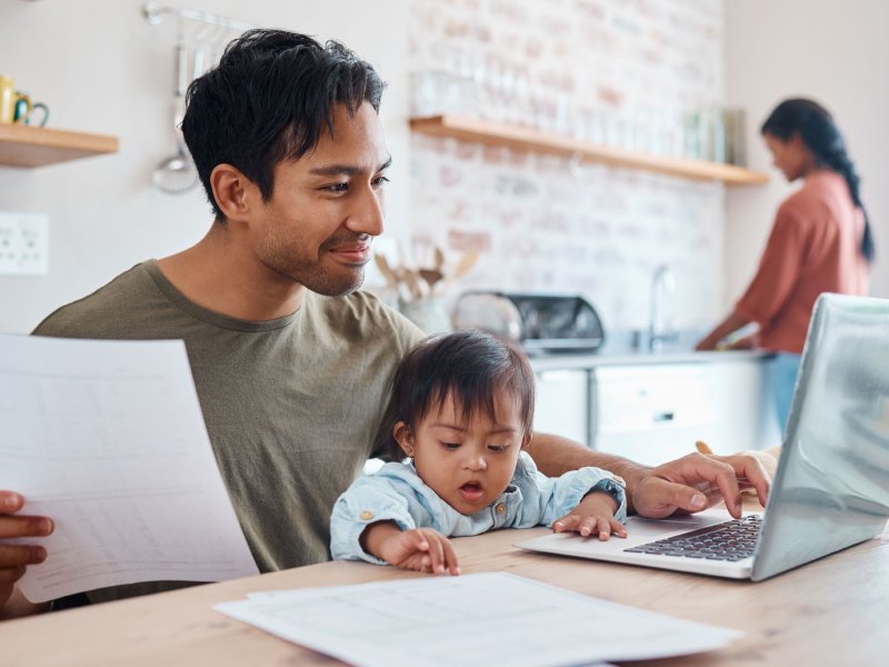 Vater sitzt mit Baby auf dem Schoß vor dem Laptop in der Küche. Die Frau steht im Hintergrund an der Spüle.