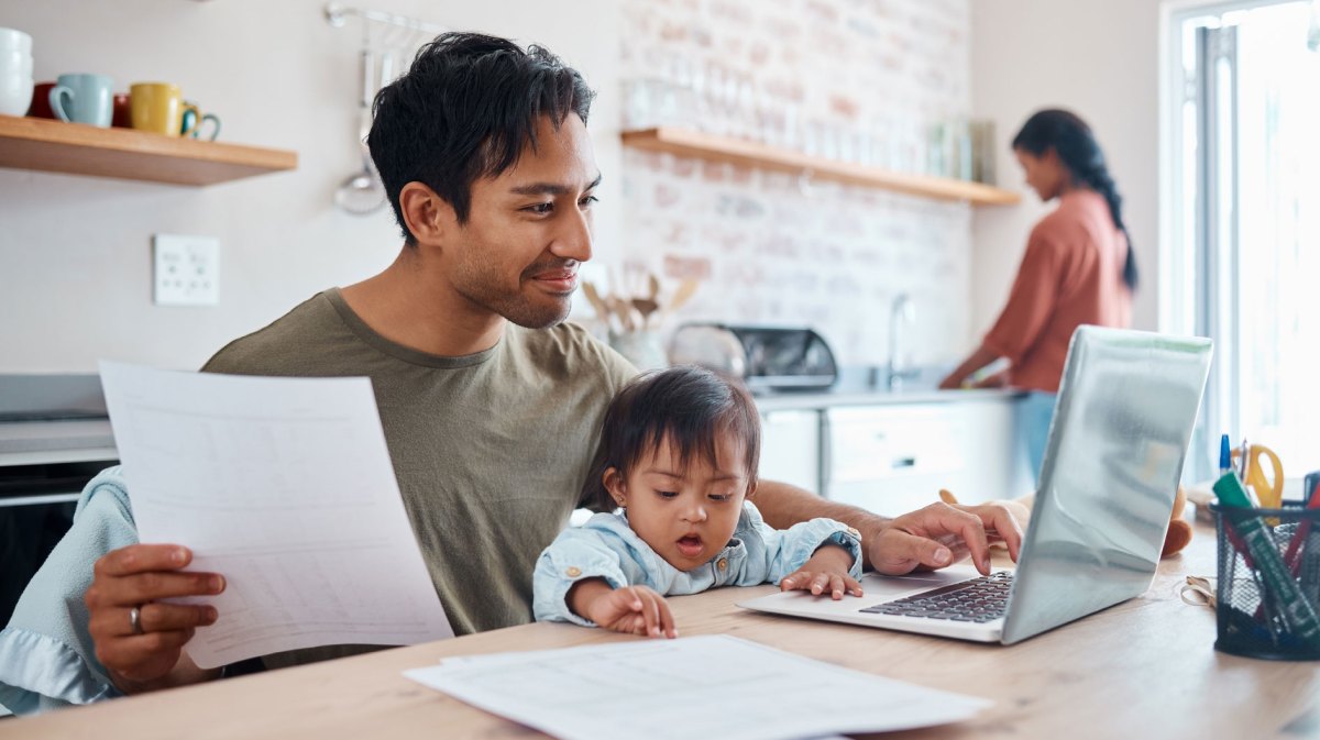 Vater sitzt mit Baby auf dem Schoß vor dem Laptop in der Küche. Die Frau steht im Hintergrund an der Spüle.