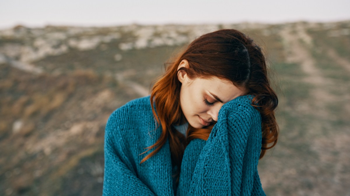 Frau mit blauem Pullover, die draußen steht und sich die Hand vor den Kopf hält