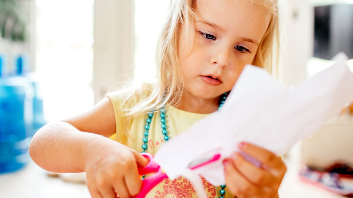 Mädchen im Vorschulalter schneidet konzentriert mit einer Schere ein Stück weißes Papier.